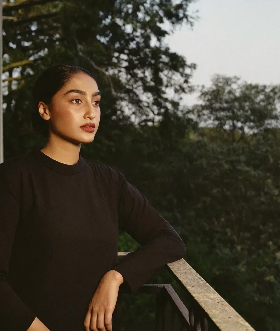 A woman in a black top leans on a balcony rail above dark trees at dusk