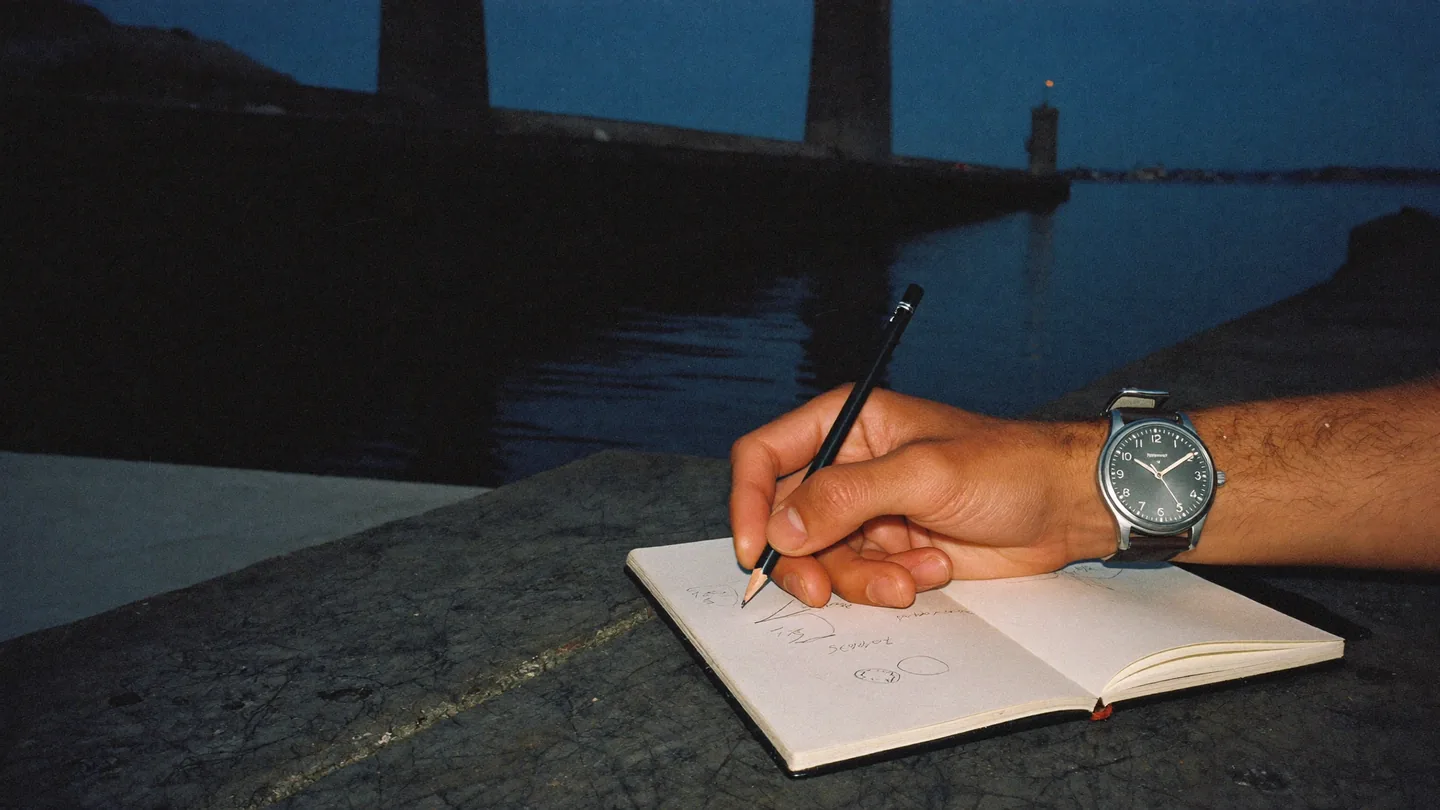 At night on a stone quay, a hand in a grey watch sketches in a notebook beside the water