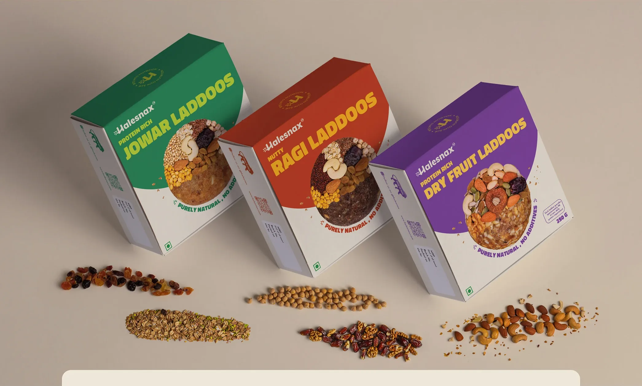 The three laddoo boxes, Jowar in green, Ragi in red and Dry Fruit in purple, angled on a beige backdrop with their ingredients scattered in front of them