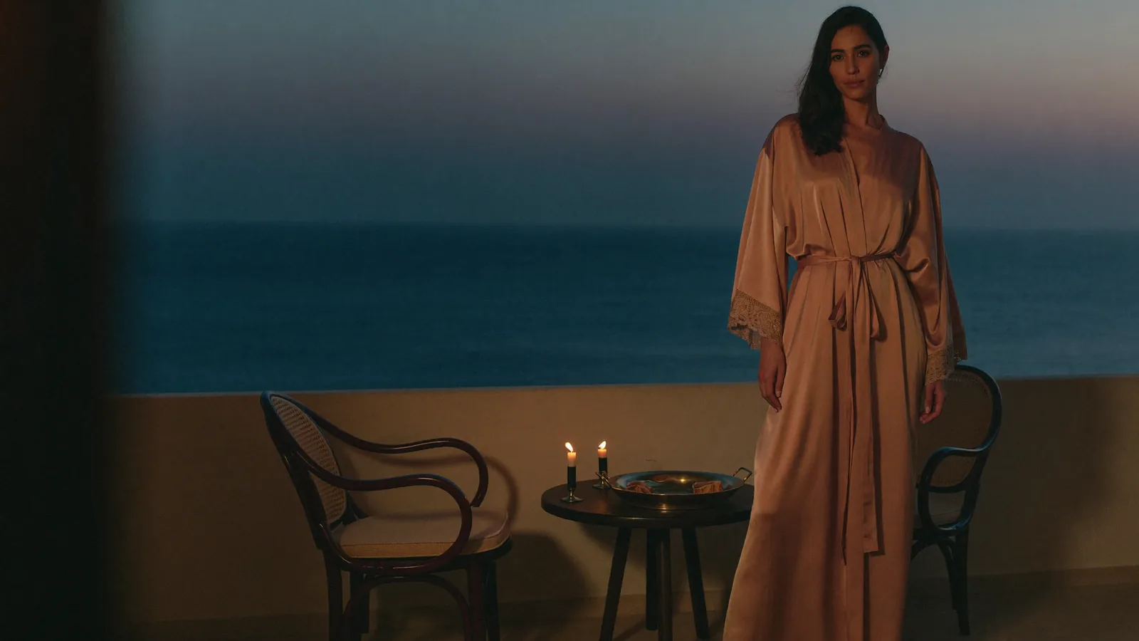 A woman in a rose silk robe on a dim sea-facing balcony, candles lit on the table beside her