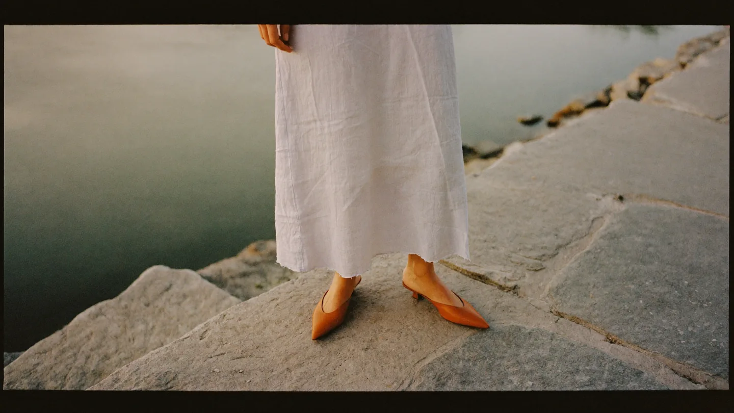 A woman in a white linen skirt and tan pointed mules, standing on stone steps by the water